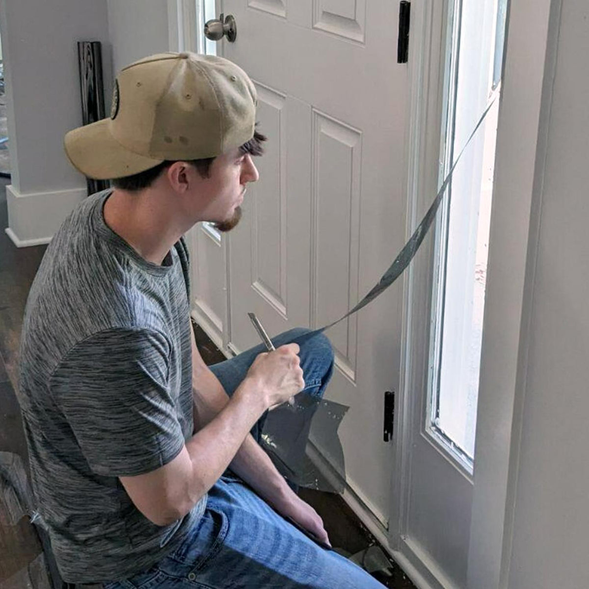A man wearing a tan baseball cap and a gray heathered t-shirt kneels on the floor next to a white paneled door, holding a long strip of window film.