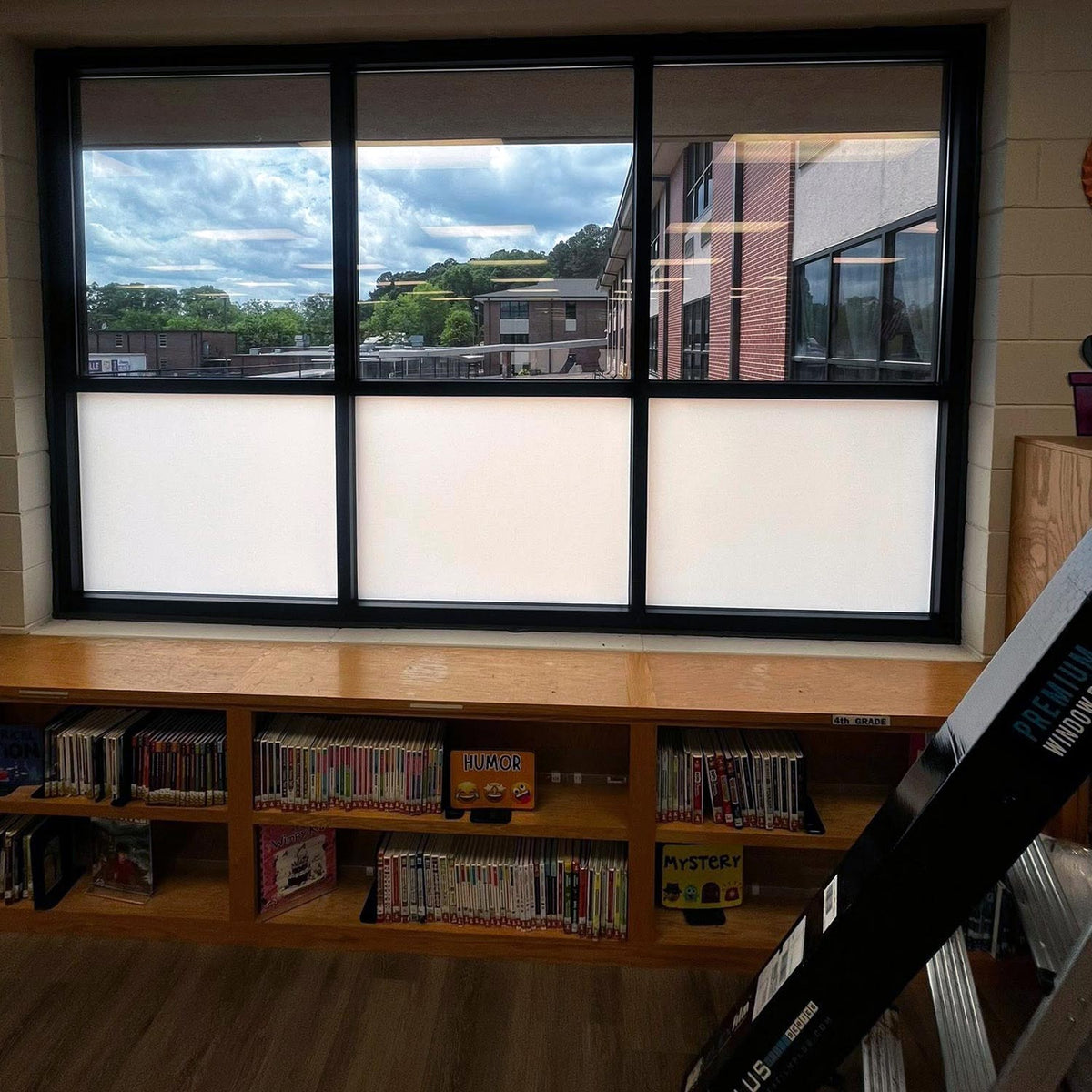 Black-framed window film installed on a multi-pane window in a school or library setting, with frosted white privacy sections covering the lower half. Bookshelves with DVDs and a ladder are visible in the room.